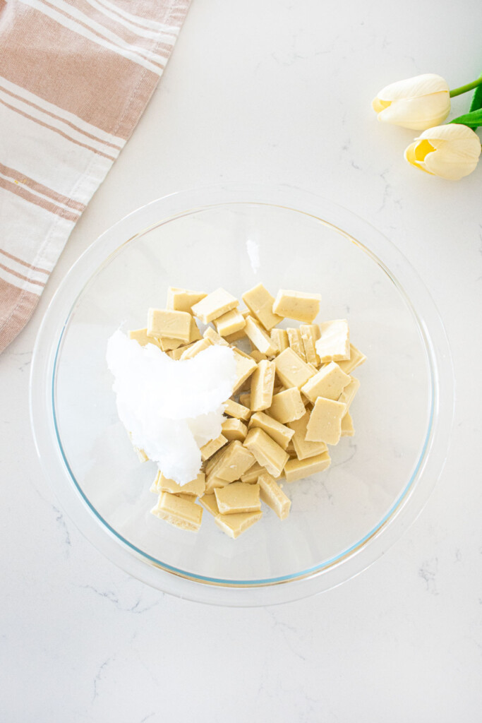 glass bowl with chopped white chocolate and coconut oil on a marble counter.