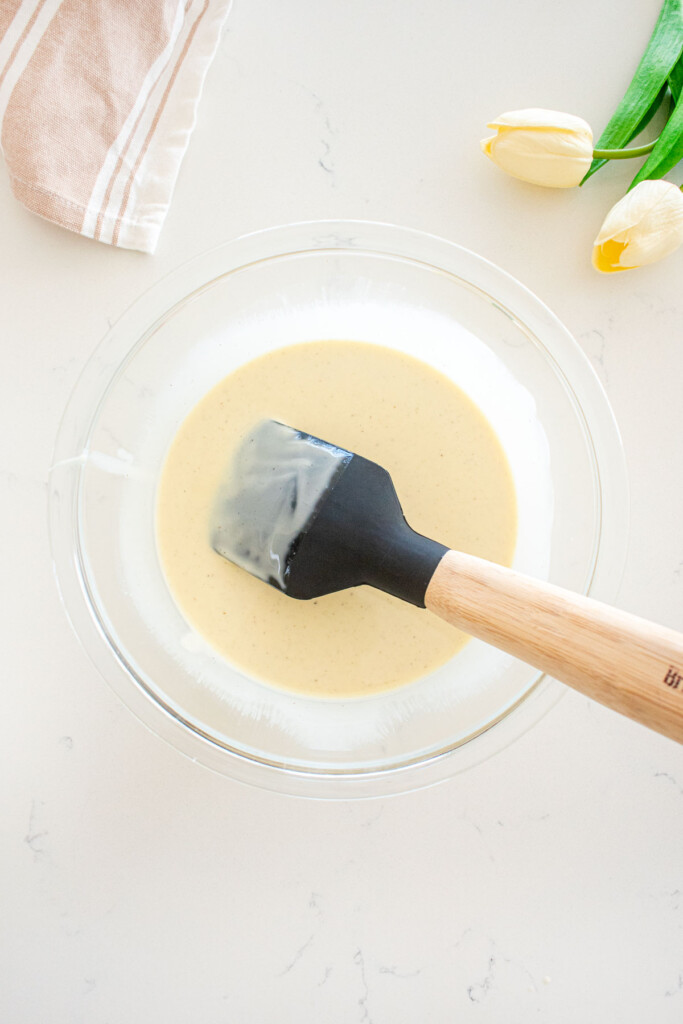 melted white chocolate and coconut oil in a glass mixing bowl with a spatula on a white marble counter.