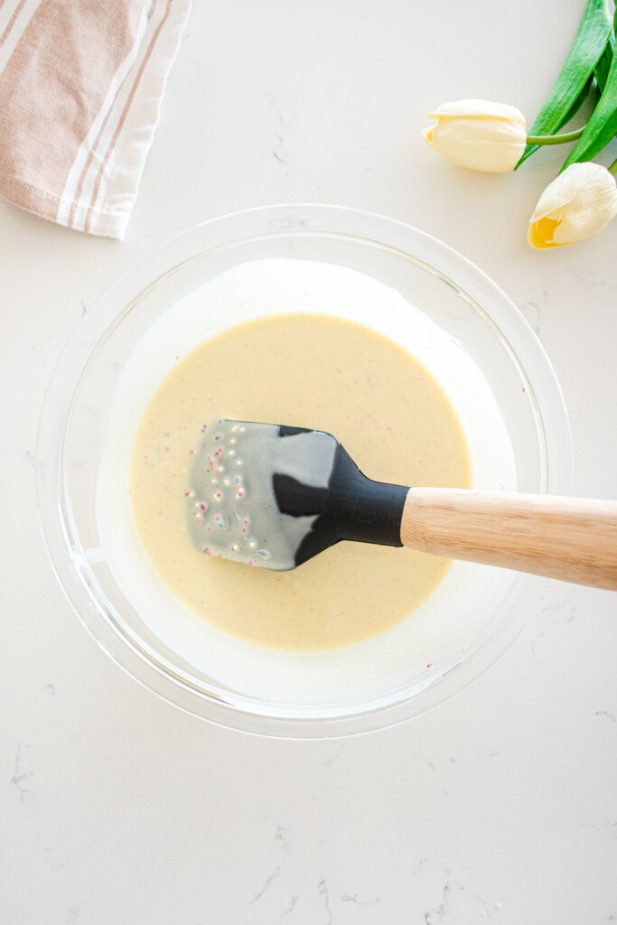 funfetti magic shell in a glass mixing bowl with a spatula on a marble counter.