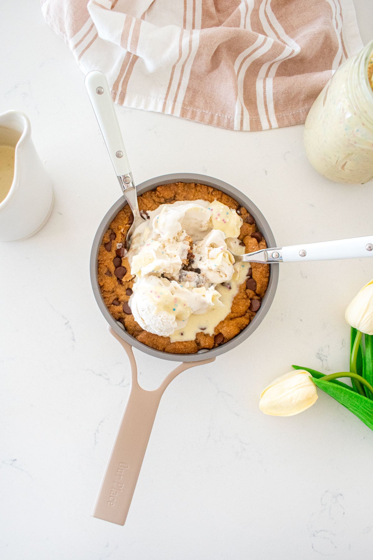 tiny chocolate chip cookie skillet with vanilla ice cream and funfetti magic shell with two spoons it in on a marble counter.