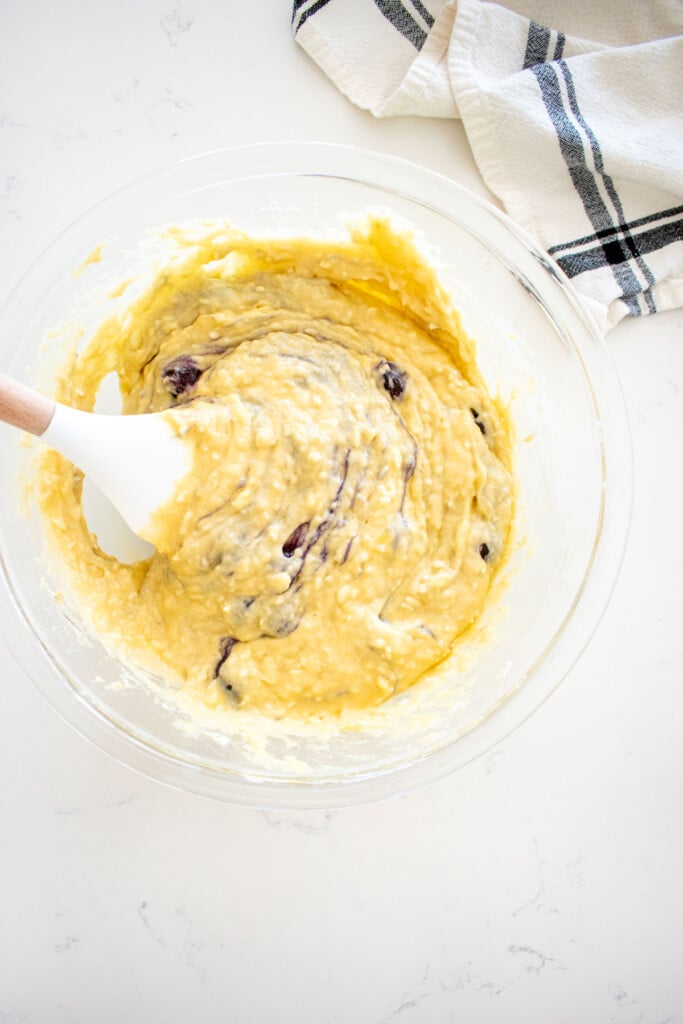 dog banana cake being stirred with blueberries in a glass mixing bowl on a marble counter.