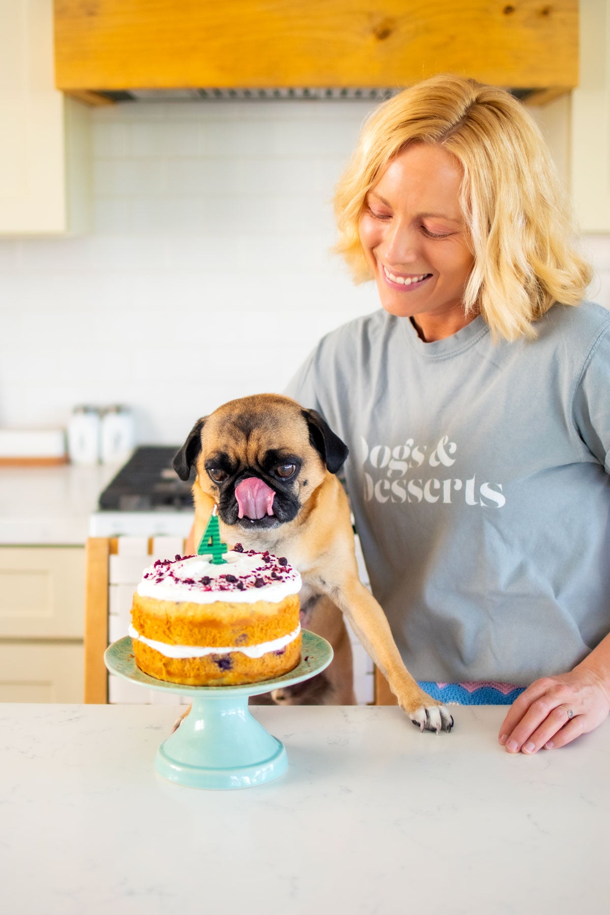 woman and pug in the kitchen with a banana blueberry dog birthday cake on a cake pedestal.