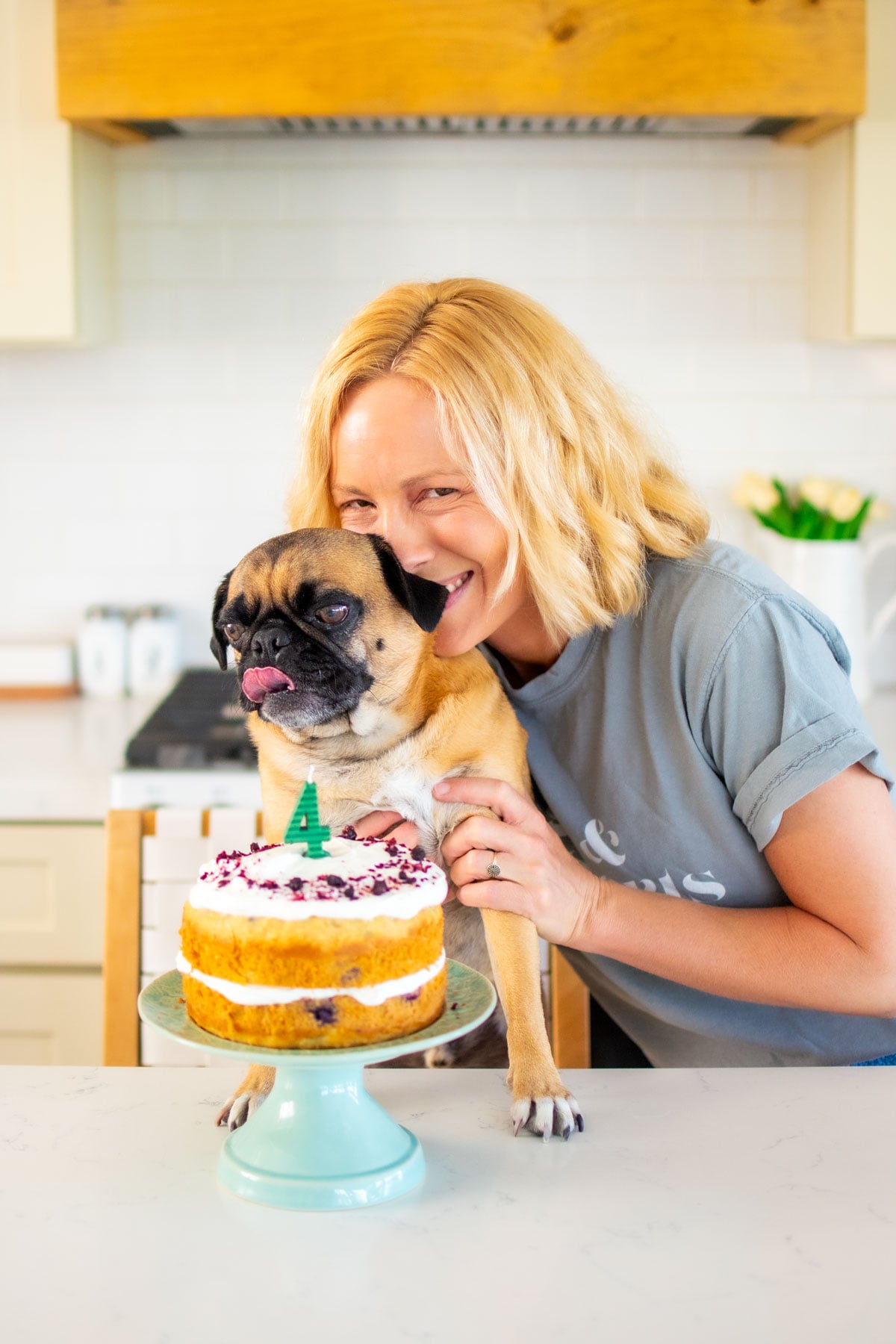 woman and pug in the kitchen with a banana blueberry dog birthday cake on a cake pedestal.