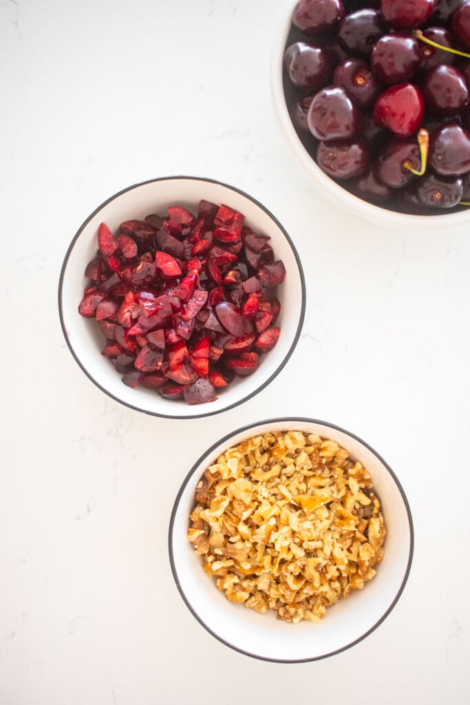bowl of diced cherries and bowl of chopped walnuts on a marble conter.