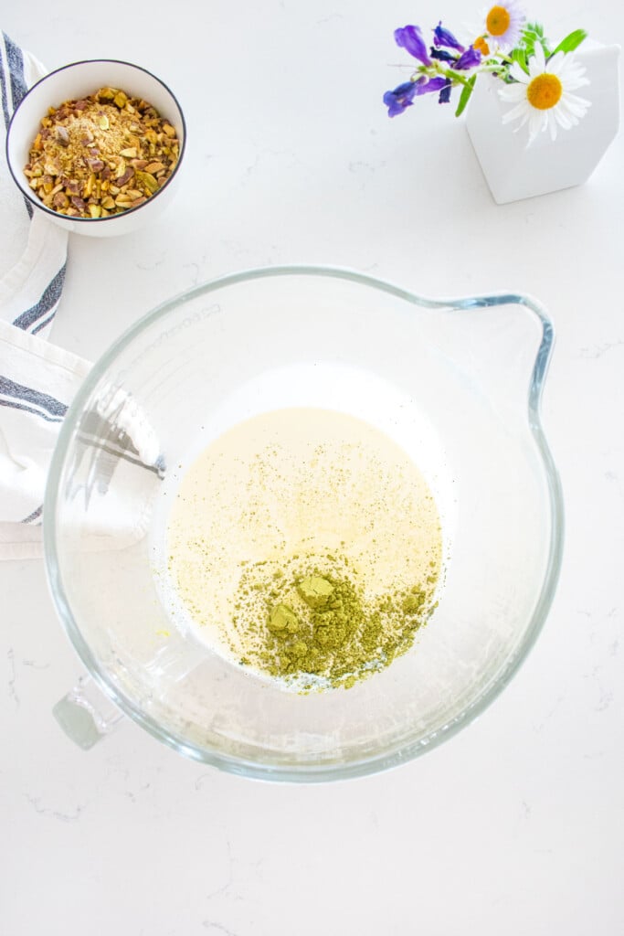 glass mixing bowl with heavy whipping cream, matcha powder and pistachio extract in it sitting on a marble counter with a small bowl of chopped pistachios next to it.