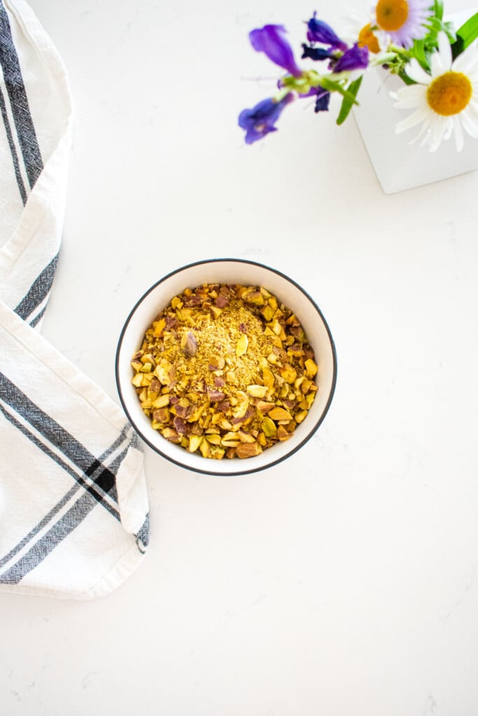 bowl of chopped pistachios on a marble counter with a small vase of flowers.