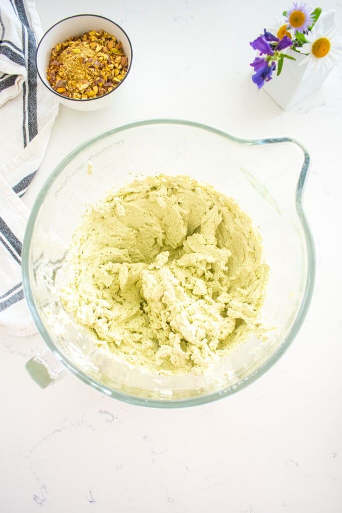 pistachio matcha whipped cream in a glass stand mixing bowl on a marble counter with a small bowl of chopped pistachios next to it.