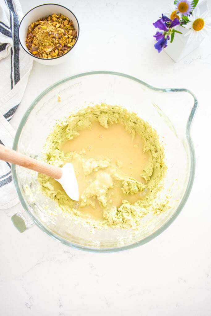 pistachio matcha whipped cream in a glass mixing bowl with sweetened condense milk added to it on a marble counter with a bowl of chopped pistachios next to it.
