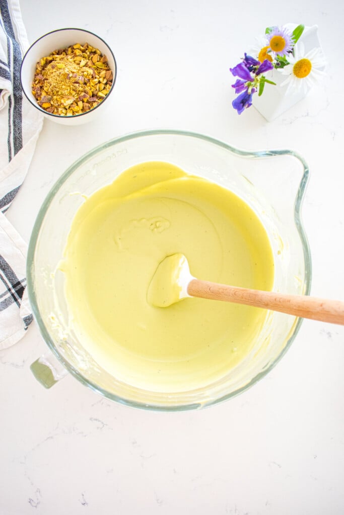 pistachio matcha ice cream base in a glass stand mixing bowl with a spatula on a marble counter with a bowl of chopped pistachios beside it.