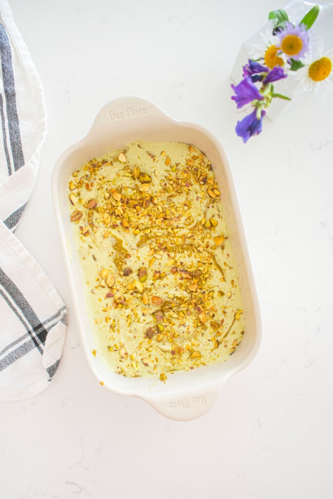 pistachio matcha ice cream in a loaf pan with swirls of pistachio butter and chopped pistachios sitting on a marble counter with fresh flowers.