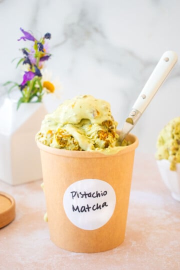 kraft ice cream container with a pistachio matcha ice cream label on it and pistachio matcha ice cream overflowing the top with a spoon on a table with a vase of flowers.