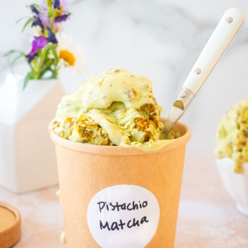 kraft ice cream container with a pistachio matcha ice cream label on it and pistachio matcha ice cream overflowing the top with a spoon on a table with a vase of flowers.