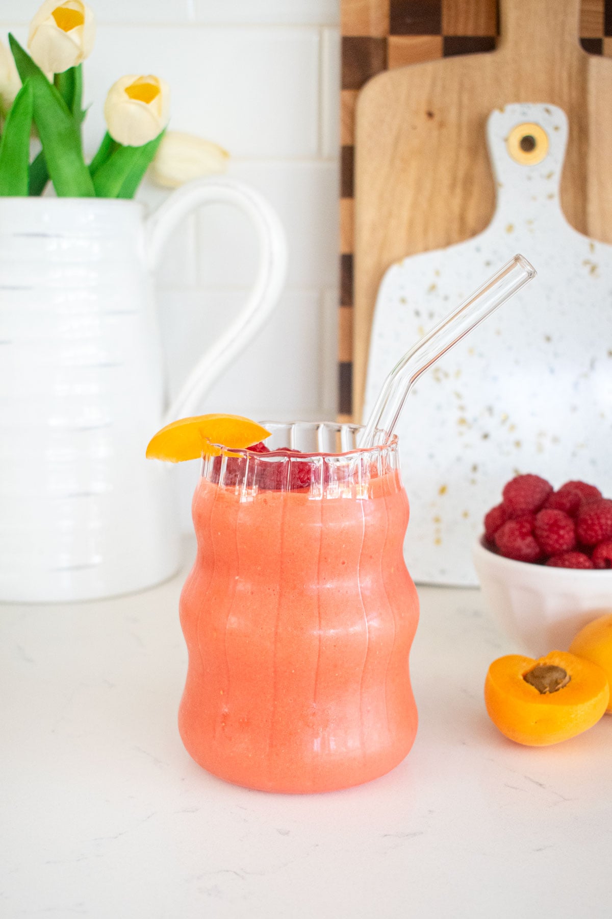 raspberry apricot smoothie in a glass with a glass straw and a fresh apricot slice on a marble counter with fresh flower vase.