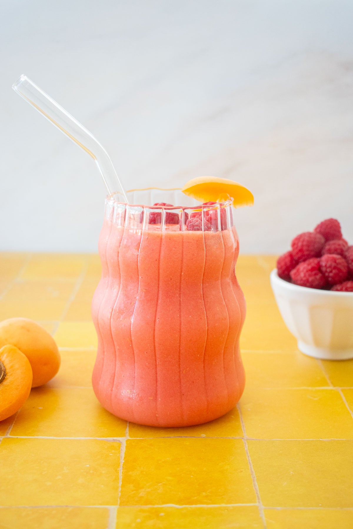bright pink raspberry apricot smoothie in a glass with a glass straw and fresh apricot slice on a tile table.