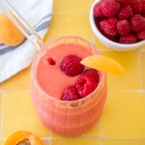 raspberry apricot smoothie in a glass with a slice of apricot and a few raspberries in the glass on a tile table.