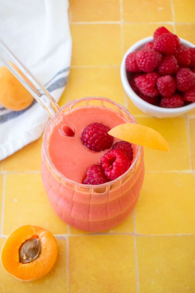 raspberry apricot smoothie in a glass with a slice of apricot and a few raspberries in the glass on a tile table.