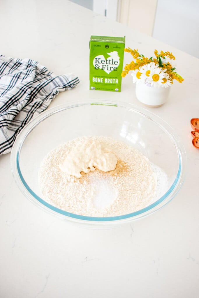 ingredients for sourdough discard bread with bone broth in a glass mixing bowl on a marble counter with a vase of flowers.