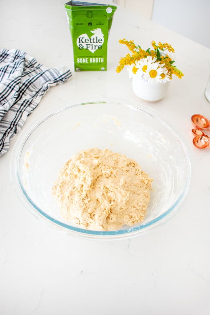 shaggy sourdough bread dough in a glass mixing bowl on a marble counter with a carton of bone broth.