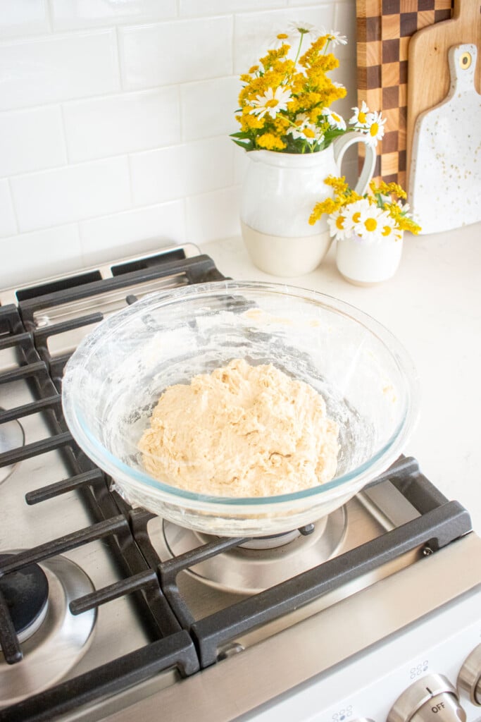 shaggy sourdough bread dough in a glass mixing bowl wrapped with plastic wrap sitting on top of an oven.