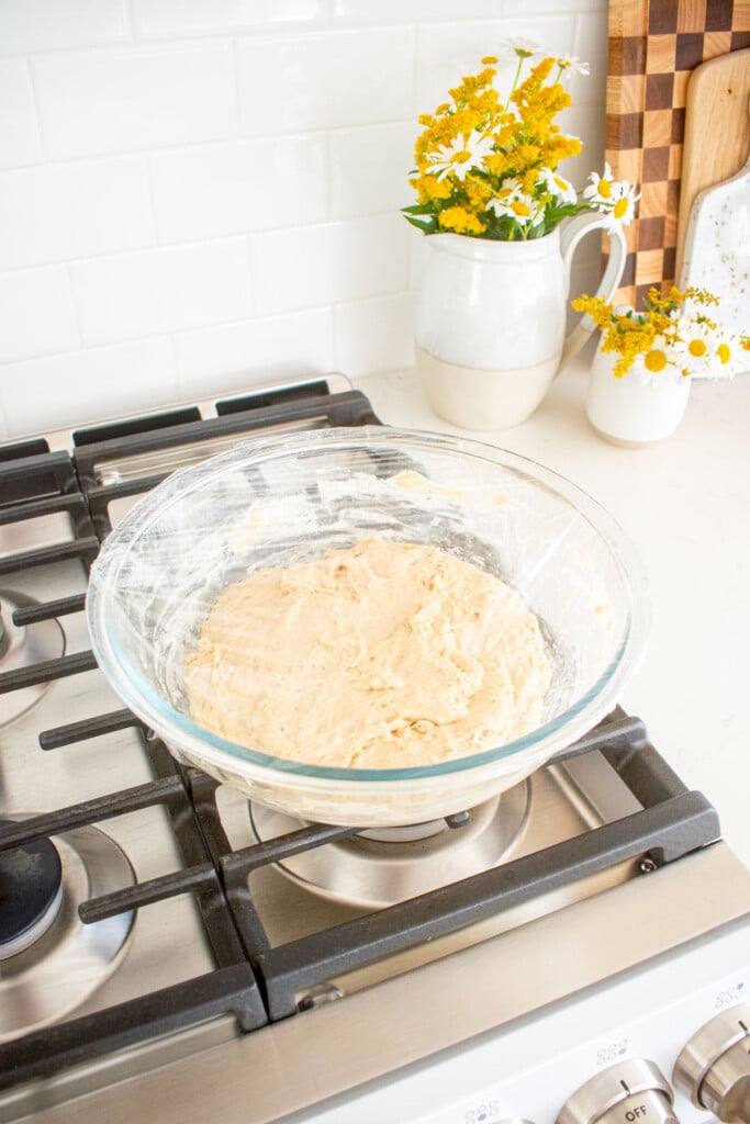 shaggy sourdough bread dough in a glass mixing bowl wrapped with plastic wrap sitting on top of an oven.