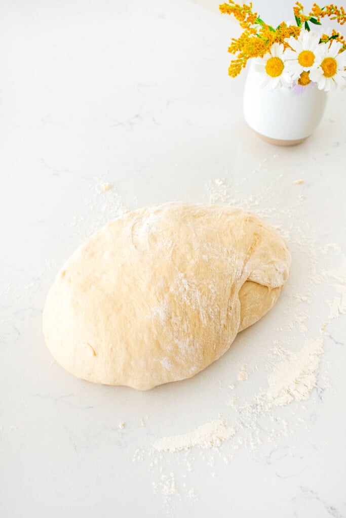 sourdough bread dough on a floured marble countertop formed into a loaf next to a small vase of flowers.