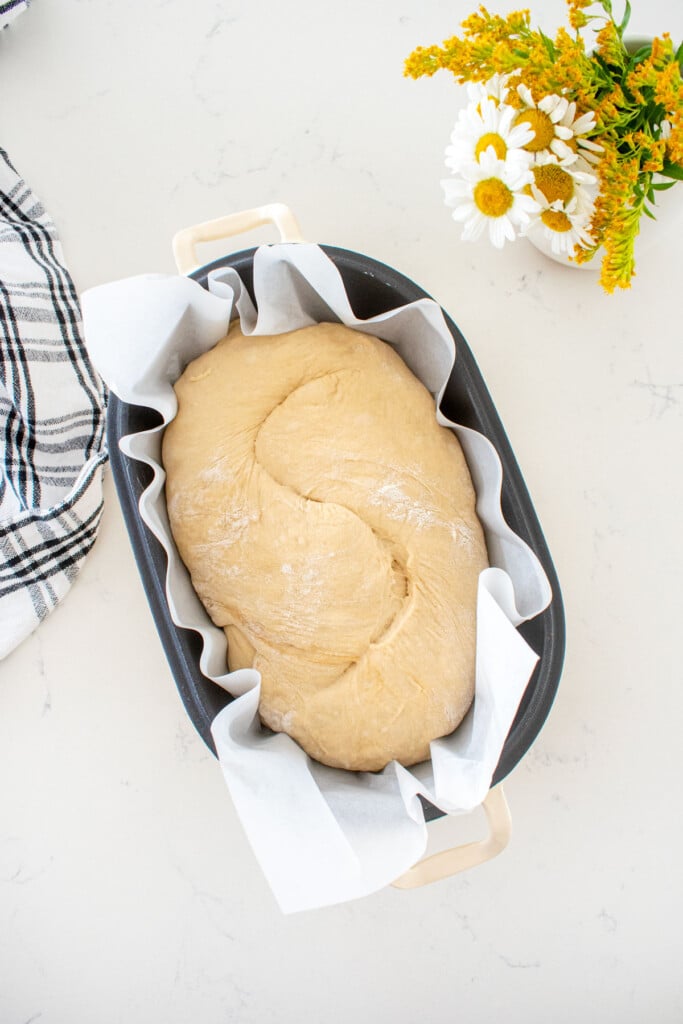 sourdough bread dough in a bread oval, scored down the center sitting on a marble counter with a small vase of flowers.