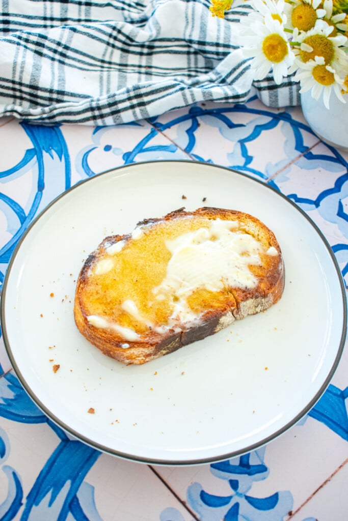 sourdough bread slathered with warm, melty butter on a plate on a tile table with fresh flowers.