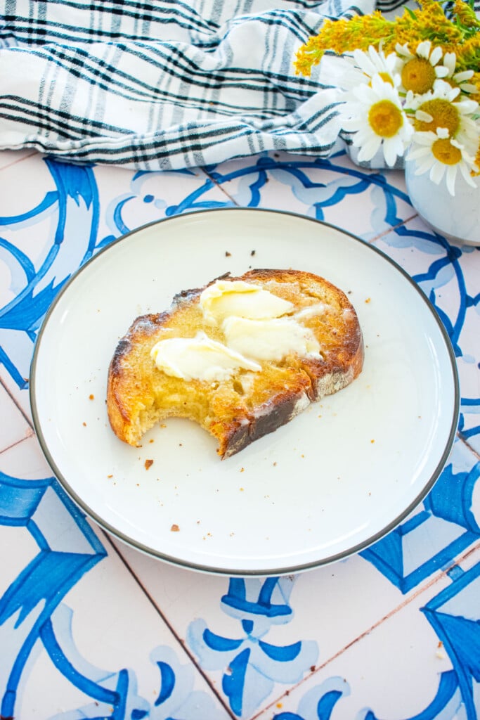 sourdough bread slathered with warm, melty butter on a plate with a bite taken out on a tile table with fresh flowers.