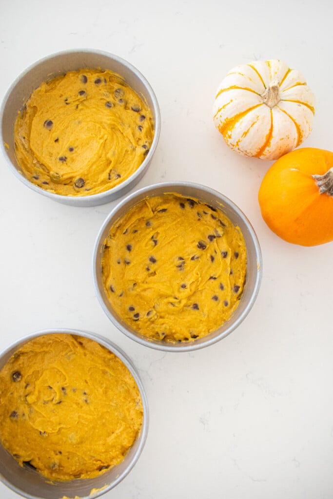 pumpkin chocolate chip cake batter in three cake pans on a marble counter with tiny pumpkins.