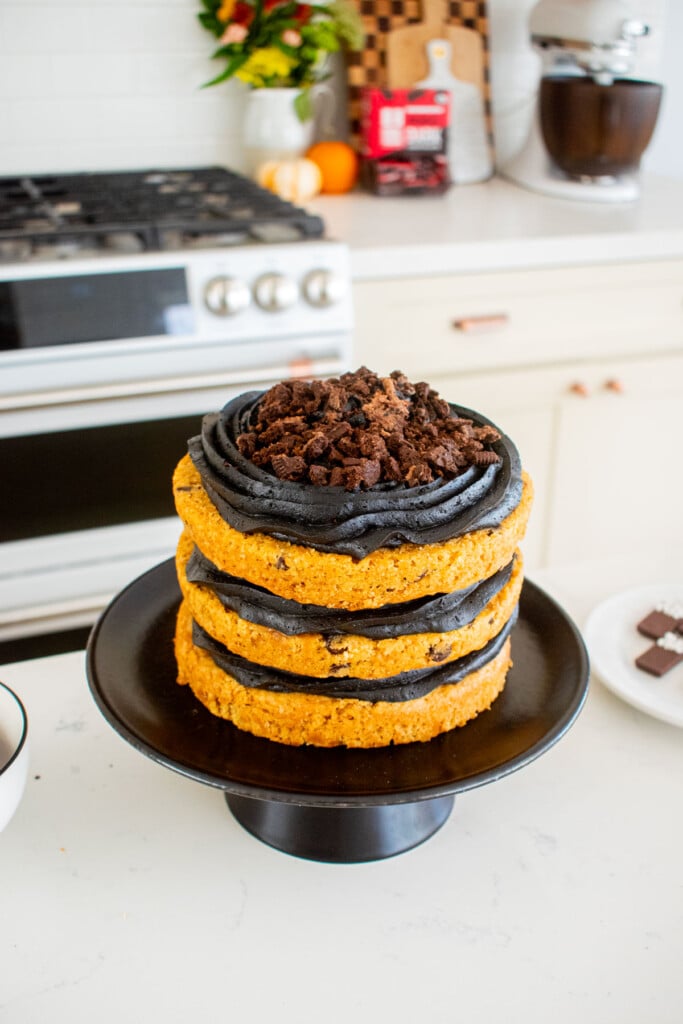 pumpkin chocolate chip naked cake with black frosting on a cake stand on kitchen counter with crumbled chocolate cookies on top.