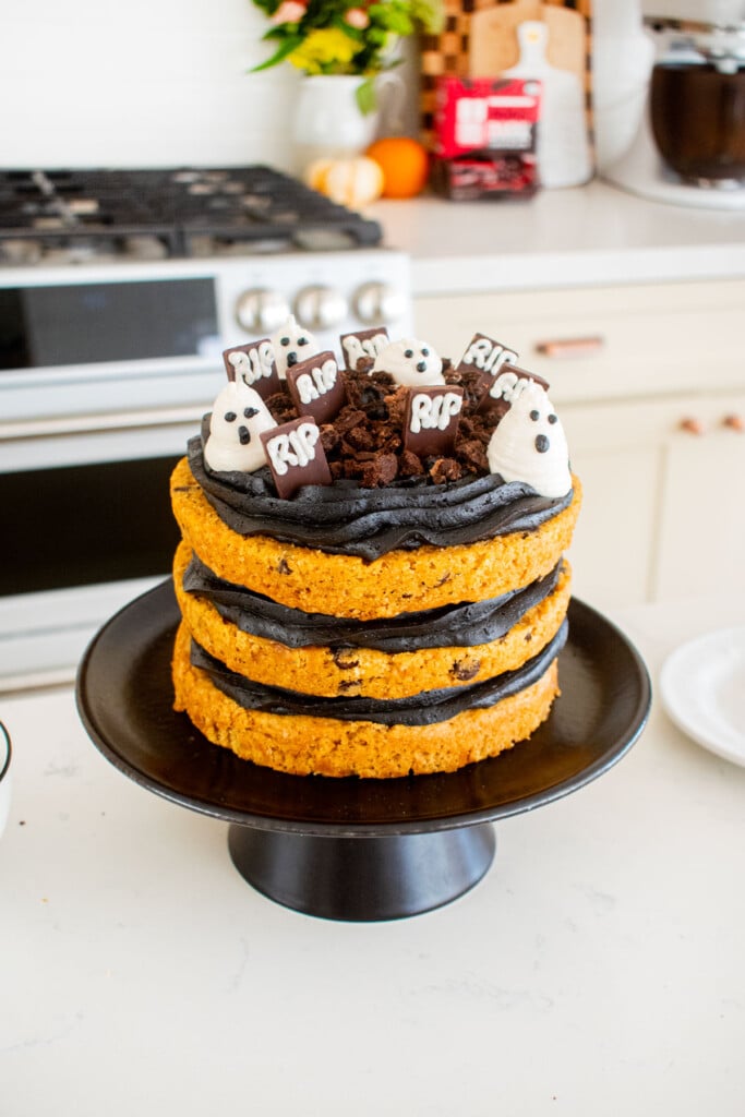 pumpkin chocolate chip naked cake with black frosting and decorated with frosting ghosts and headstones like a graveyard on a cake stand on kitchen counter.