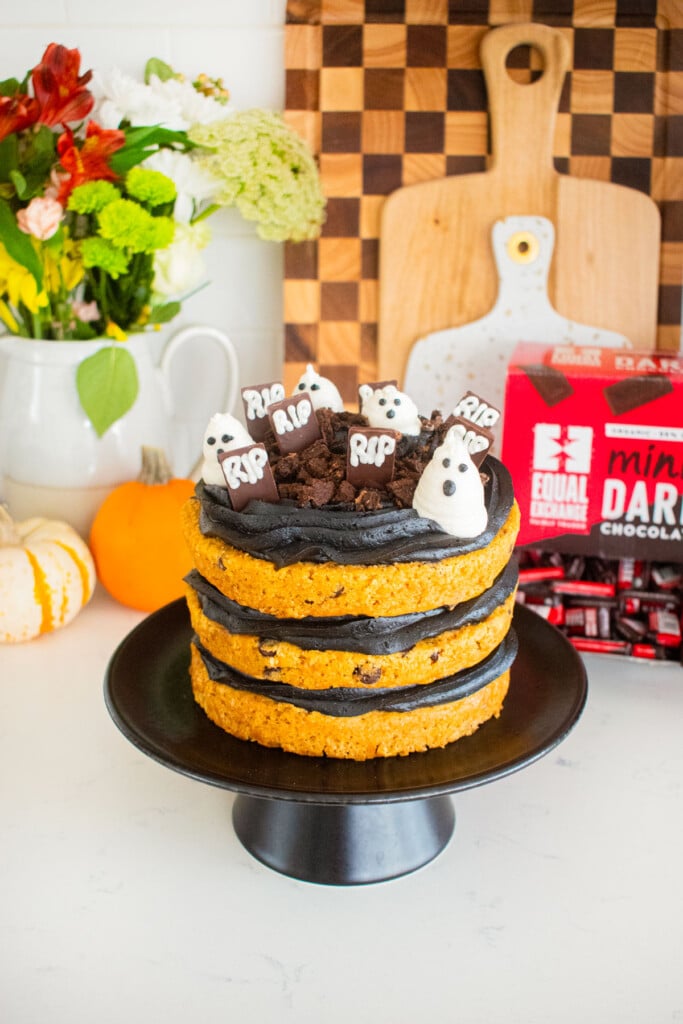 pumpkin chocolate chip naked cake with black frosting and a graveyard design on top sitting on a marble counter with tiny pumpkins and fresh flowers.