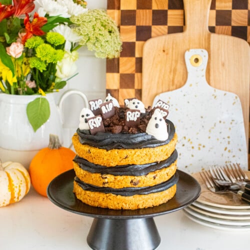 pumpkin chocolate chip naked cake with black frosting and a graveyard design on top sitting on a marble counter with tiny pumpkins and fresh flowers.