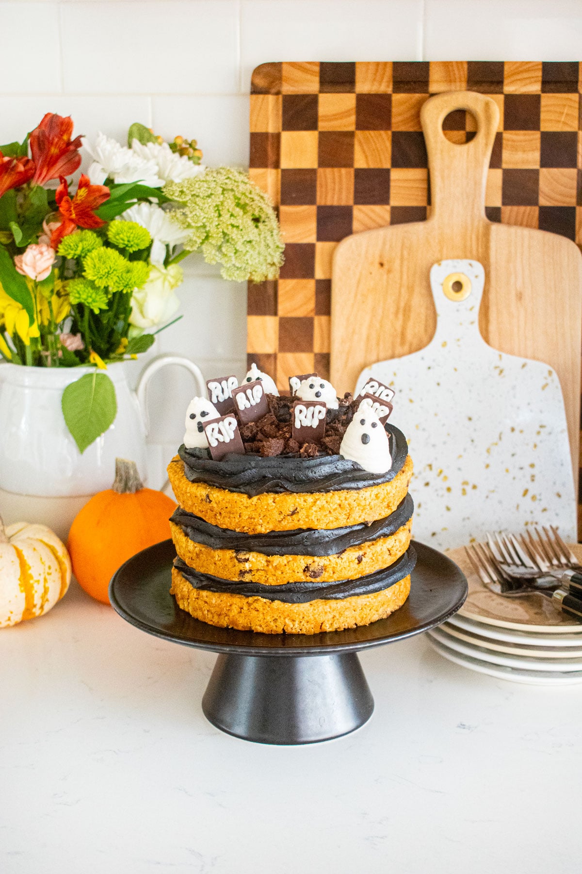 pumpkin chocolate chip naked cake with black frosting and a graveyard design on top sitting on a marble counter with tiny pumpkins and fresh flowers.