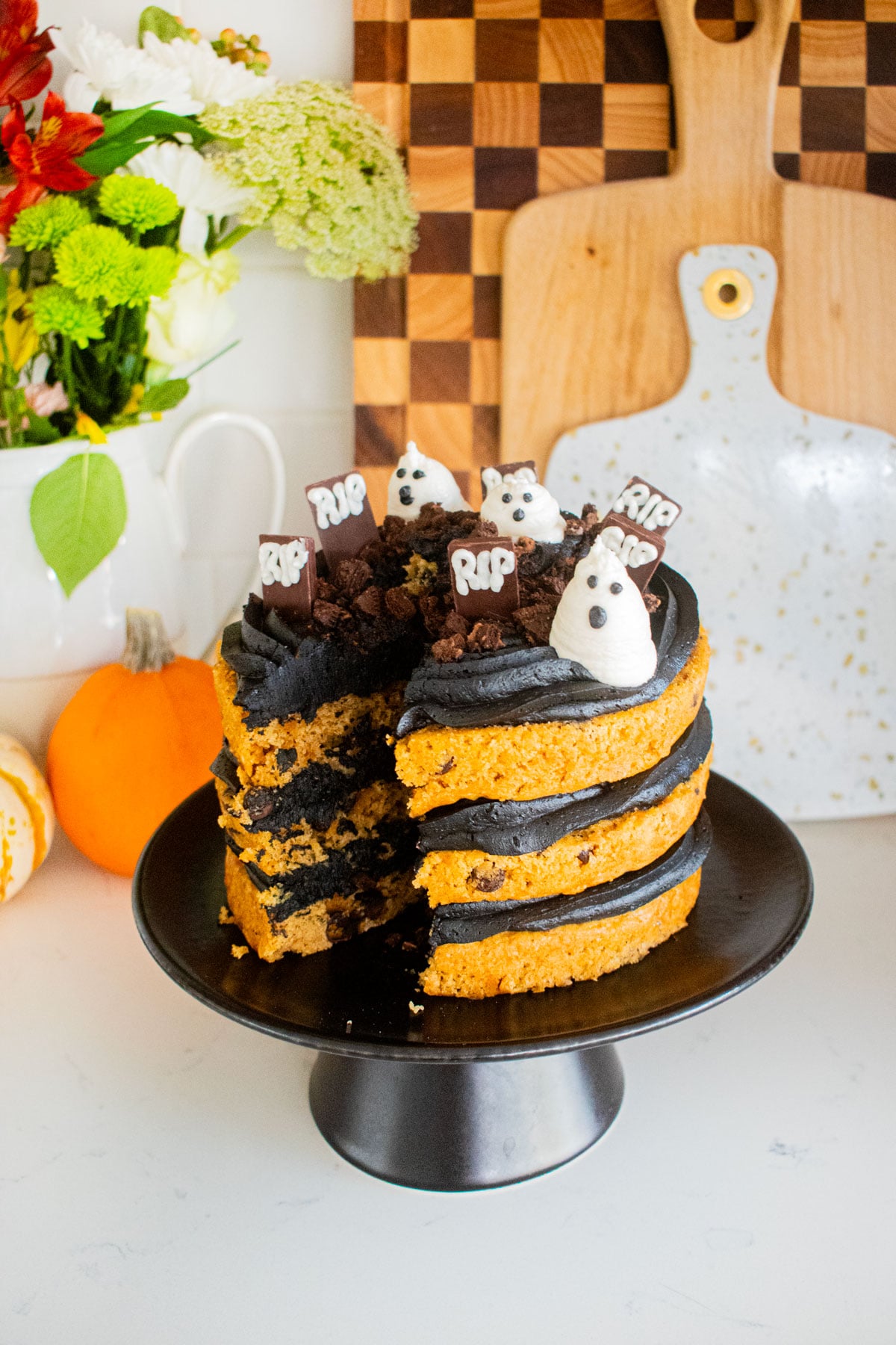 pumpkin chocolate chip naked cake with black frosting and a graveyard design on top with a slice removed sitting on a marble counter with tiny pumpkins and fresh flowers.