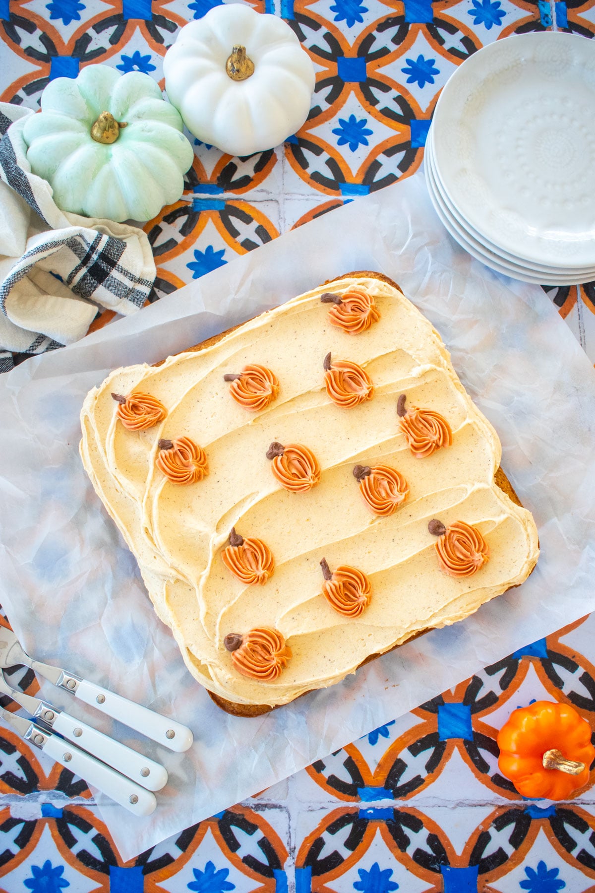 square single layer cake with pumpkin buttercream frosting on top with small piped pumpkins on a tile table with dessert plates and pumpkins.