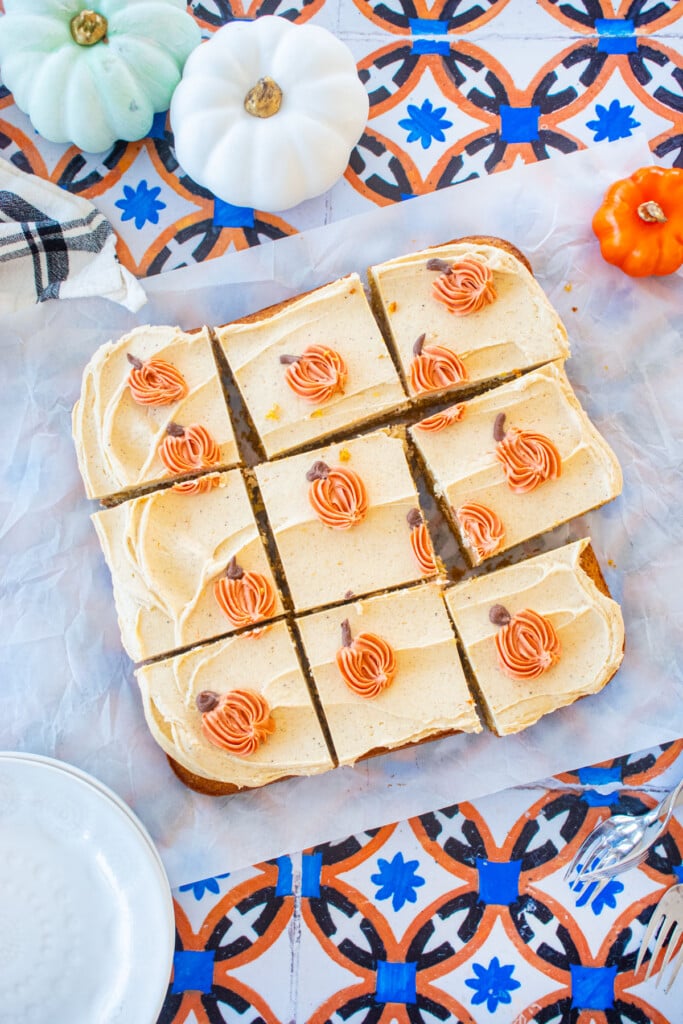 square single layer cake with pumpkin buttercream frosting on top with small piped pumpkins cut into slices on a tile table with dessert plates and pumpkins.