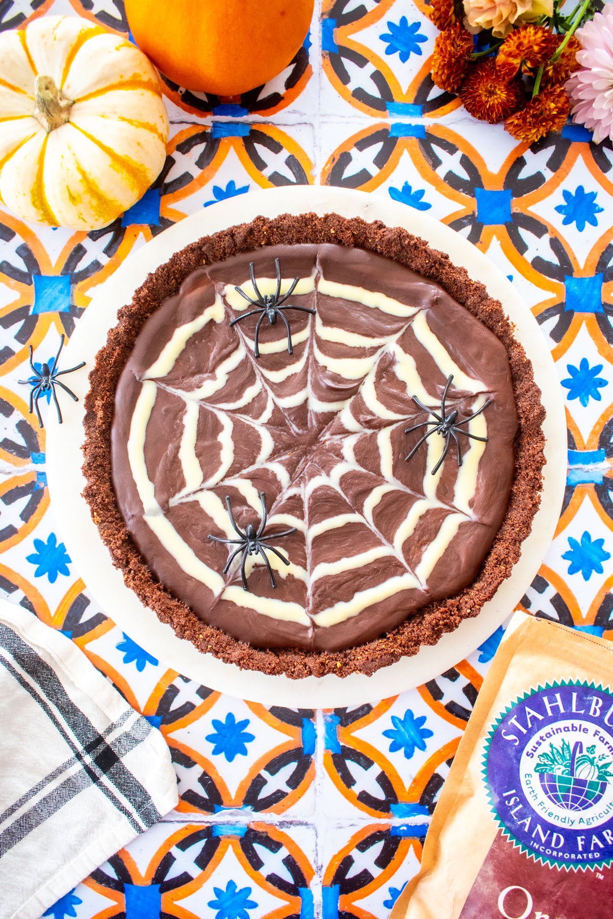 spider web chocolate strawberry tart on a marble serving board on a tile table with tiny pumpkins and flowers.