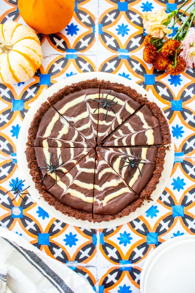 spider web chocolate strawberry tart cut into slices on a marble serving board on a tile table with tiny pumpkins and flowers.