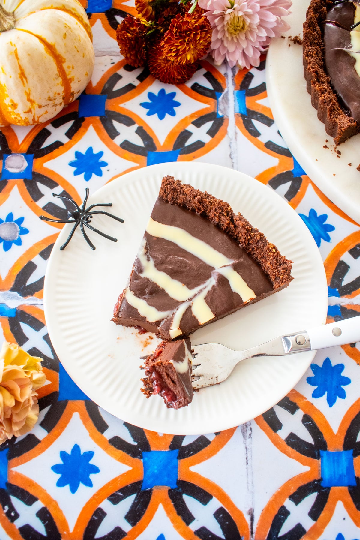 slice of spider web chocolate strawberry tart with a fork on a plate on a tile table with pumpkins and flowers.