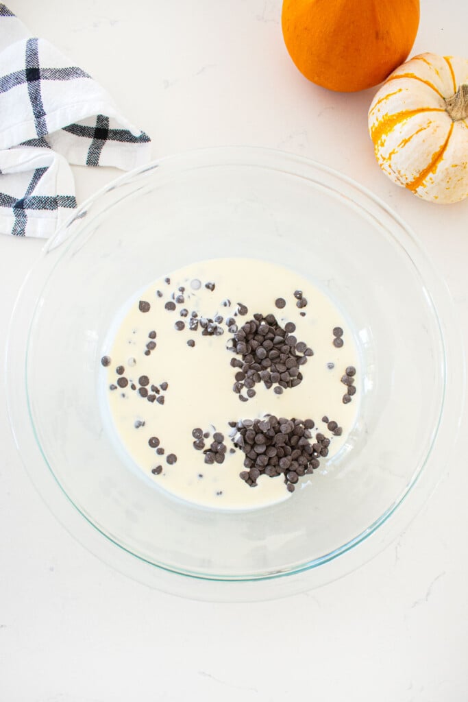 glass mixing bowl with cream and dark chocolate on a marble counter with tiny pumpkins.
