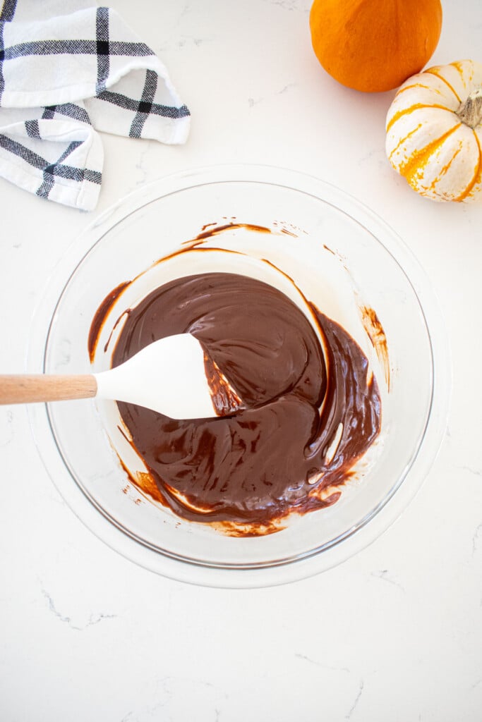 chocolate ganache in a glass mixing bowl with a spatula on a marble counter with tiny pumpkins.