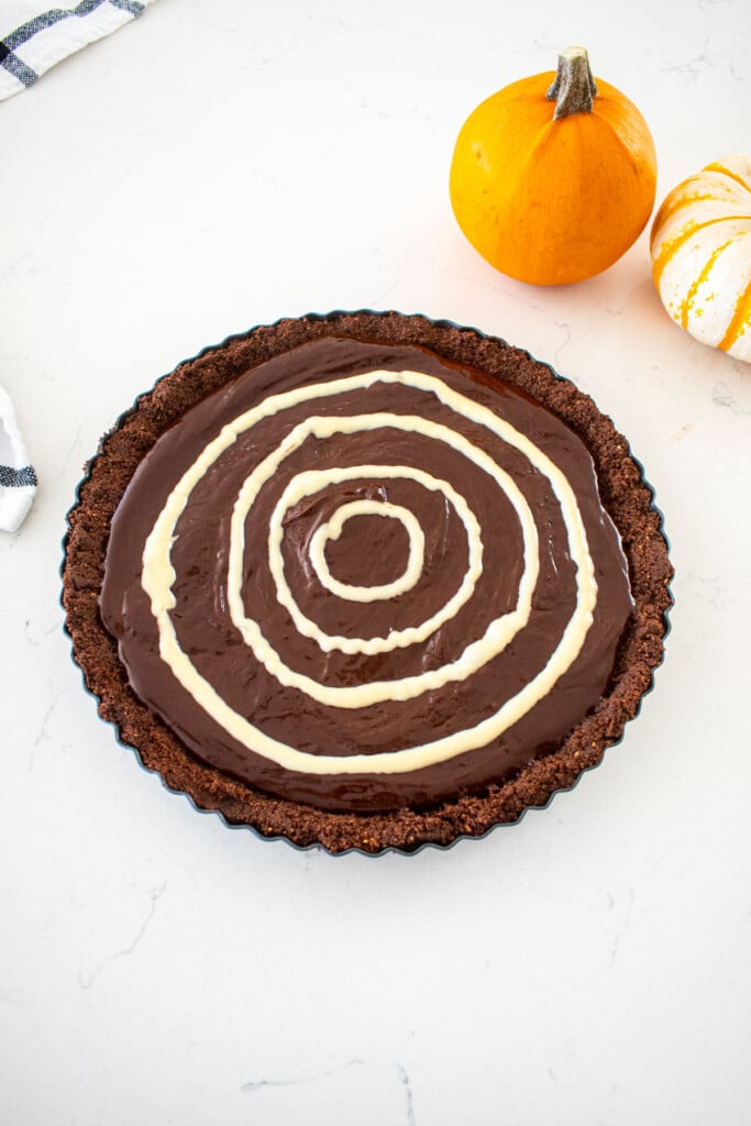 chocolate tart with white chocolate circles piped on top on a marble counter with tiny pumpkins.