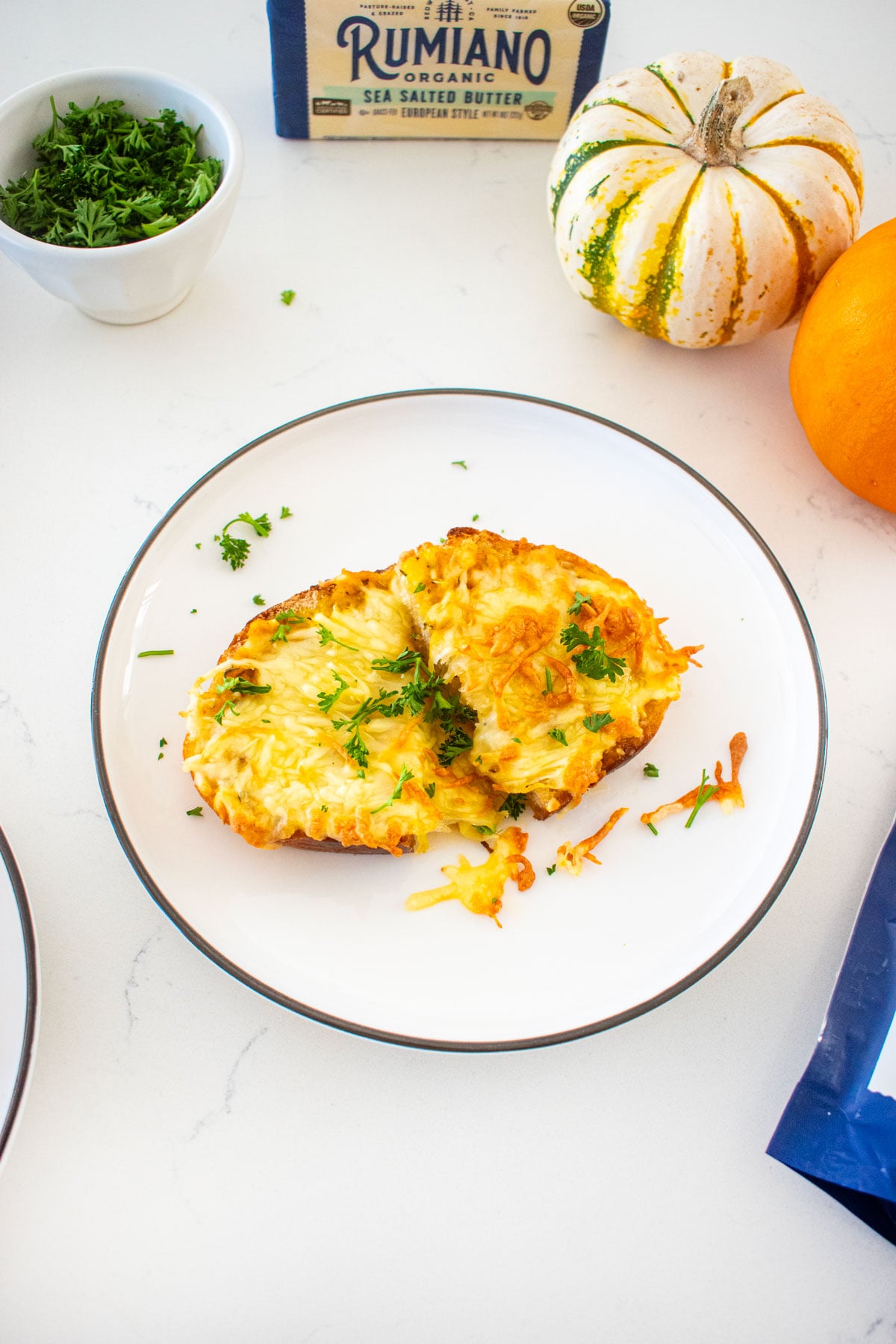 plate of cheesy pumpkin garlic bread with fresh parsley on a marble counter with tiny pumpkins and bags of shredded cheese.