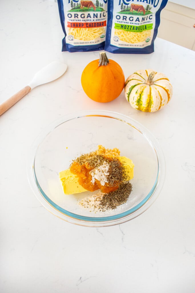 glass mixing bowl with ingredients for pumpkin garlic butter on a marble counter with tiny pumpkins and bags of shredded cheese.