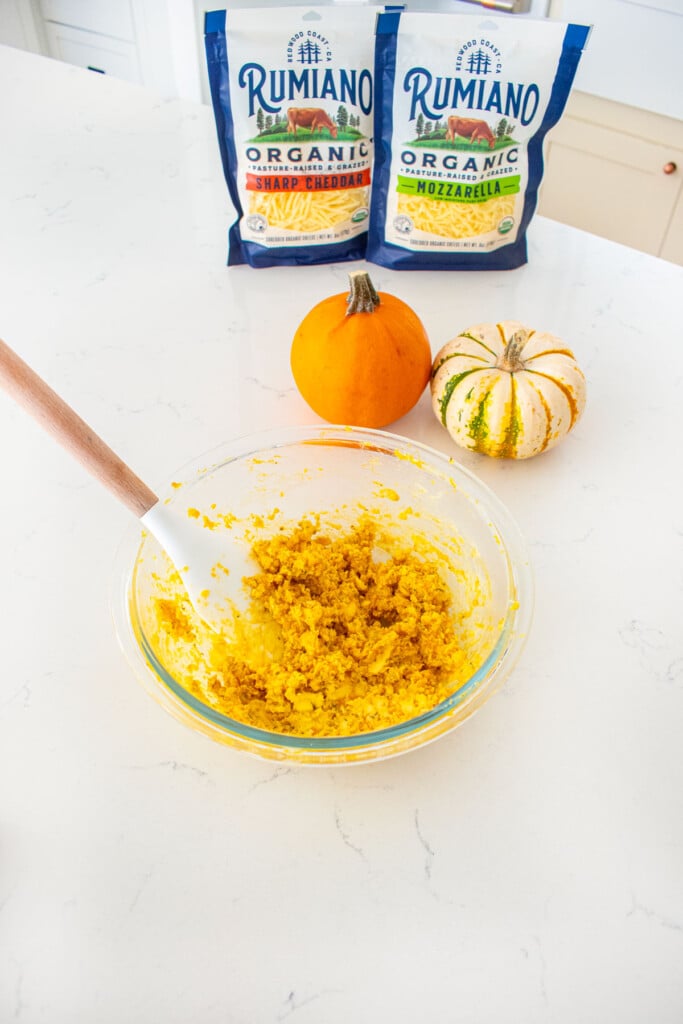 glass mixing bowl with pumpkin garlic butter mixed with a spatula on a marble counter with tiny pumpkins and bags of shredded cheese.