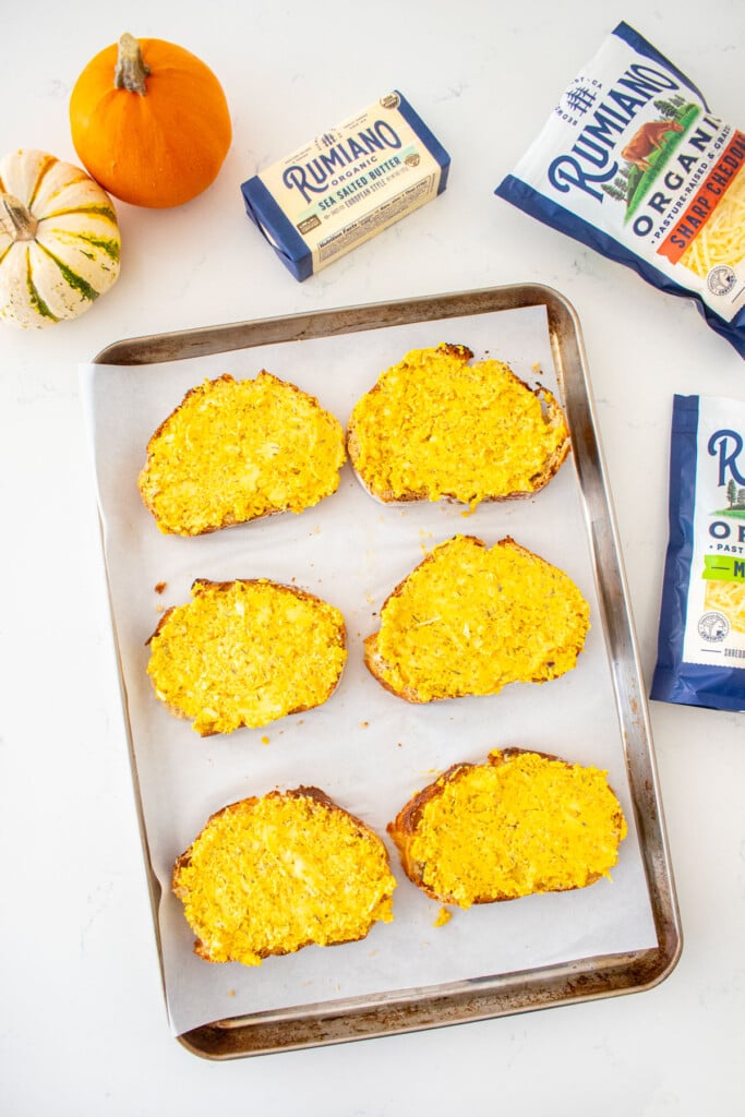 baking sheet with slices of bread spread with pumpkin garlic butter on a marble counter with tiny pumpkins and bags of shredded cheese.