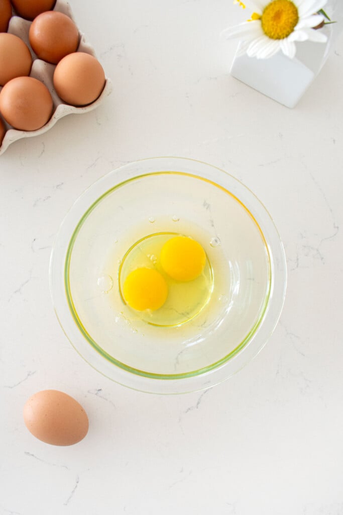 glass bowl with two eggs in it on a marble counter with a tray of brown eggs and a vase of flowers.