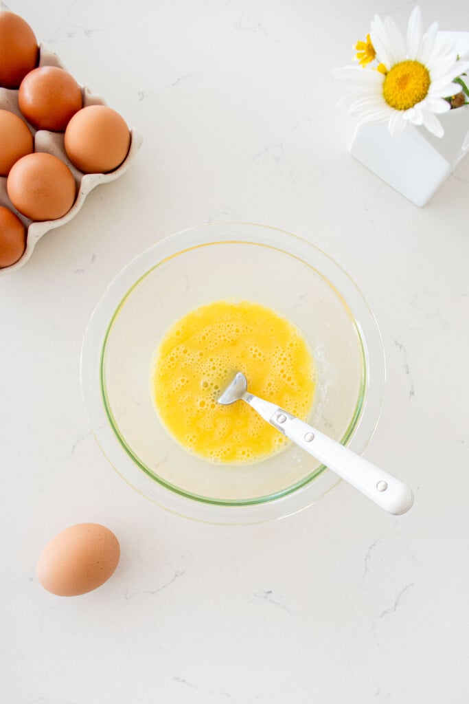 glass bowl with two eggs scrambled with a fork in it on a marble counter with a tray of brown eggs and a vase of flowers.
