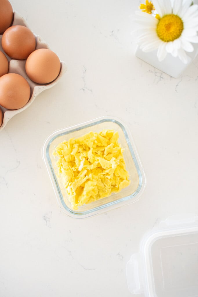 glass tupperware with scrambled eggs on a marble countertop with a tray of brown eggs and vase of flowers.